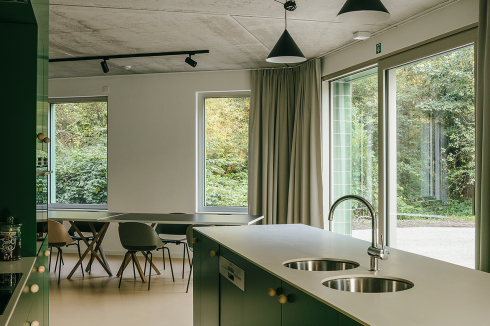 Der Gemeinschaftsraum im Erdgeschoss öffnet sich über große Fenster zur Natur. Die Bewohner und Bewohnerinnen essen und kochen gemeinsam.<br />