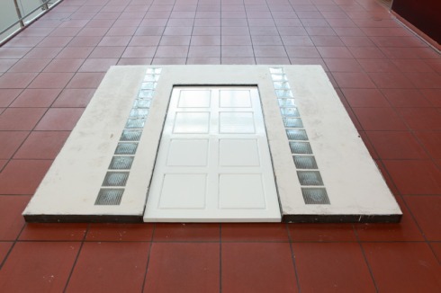 Im Kassenraum: Eine Hauseingangstür, ebenfalls um neunzig Grad auf den Boden gekippt- "Doors On The Floor", 2014; Gips, Stahl, Glass, Eichenholz, Farbe.<br />